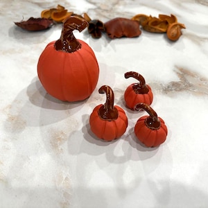 May include: A set of four orange pumpkins with dark brown stems. One large pumpkin is positioned to the left, while three smaller pumpkins of varying sizes are arranged to the right. Each pumpkin features vertical ridges and a glossy brown stem.