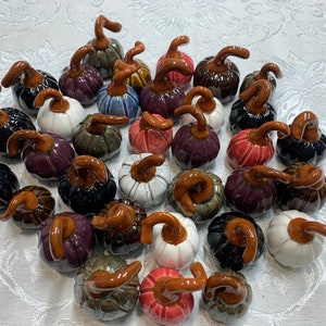 May include: A collection of ceramic pumpkins in various colors, including black, white, purple, and red. Each pumpkin has a brown, curved stem. The pumpkins are arranged on a white surface.