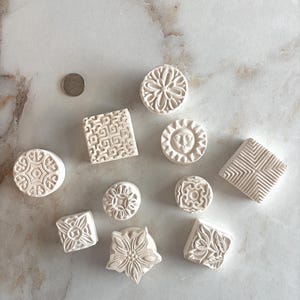 May include: A set of ten white ceramic stamps with various geometric and floral designs. The stamps are arranged on a light gray marble surface.