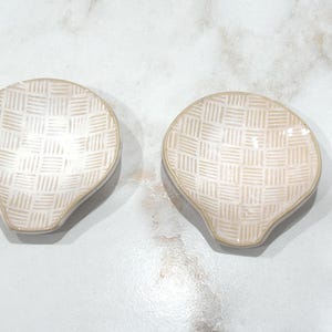 May include: Two off-white, shell-shaped dishes with a woven pattern. The dishes have a slightly irregular shape and are placed side by side. The background is a light-colored, marble-like surface.