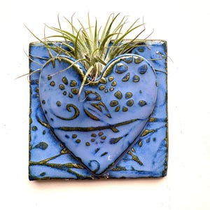 May include: A square blue ceramic wall planter featuring a raised heart-shaped pocket with a glossy finish. The surface is decorated with dark green embossed floral and bird patterns. A small green air plant sits inside the heart-shaped opening.