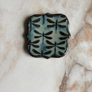 May include: A square, ceramic trinket dish with a scalloped edge. The dish features a light blue background with a repeating pattern of black dragonfly silhouettes. The dish is glazed and has a glossy finish.