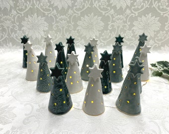 May include: A selection of ceramic Christmas tree ornaments in shades of green and white. Each tree is topped with a star and has small holes that emit a warm, yellow light. The trees are displayed on a white surface.