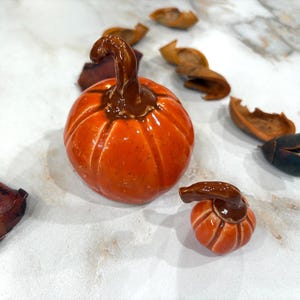 May include: Two glossy orange ceramic pumpkins with dark brown stems. The larger pumpkin features deep vertical ridges and a curved stem, while the smaller pumpkin has a similar rounded shape and a dark brown stem, both with a smooth finish.