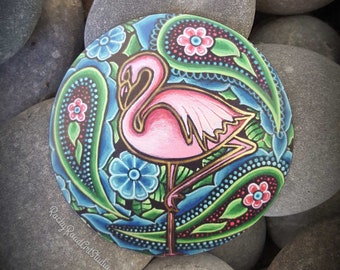 May include: A hand-painted, round stone featuring a pink flamingo with gold accents, set against a backdrop of blue and green paisley patterns and flowers. The artwork is displayed on a bed of gray river stones. The artist's signature is visible.