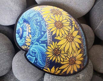 May include: A sticker with a black border featuring a design of sunflowers and paisley patterns. The sunflowers are yellow with brown centers, set against a dark blue background. The paisley section is light blue on a dark blue background.