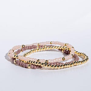 Puede incluir: Tres pulseras de cuentas en tonos rosa, malva y dorado. Las pulseras están hechas de pequeñas cuentas redondas y detalles dorados. Las pulseras están apiladas sobre una superficie blanca.