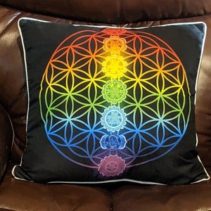 May include: A black pillow with a white border features a colorful mandala design with a flower of life pattern and a vertical line of seven chakras.