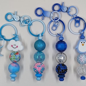 May include: Three blue and white keychains with different charms. The charms are a cloud with a smiley face, a penguin, and a sparkly blue bead. Each keychain has a silver metal ring and a blue metal clasp.