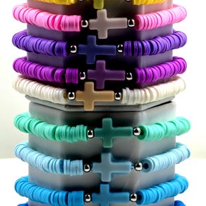 May include: Stack of colorful bracelets with cross charms. The bracelets are made of small, flat, round beads in shades of yellow, pink, purple, white, green, and blue. Each bracelet has a matching cross charm and silver-colored beads.