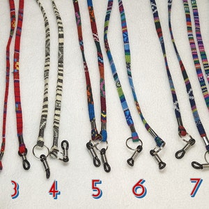May include: Nine colorful eyeglass chains with black metal clasps. The chains are made of woven fabric with various patterns and colors, including red, blue, green, yellow, and white.