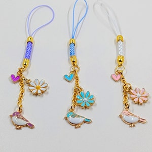May include: Three phone charms with gold chains and colorful enamel charms. Each charm features a bird, a flower, and a heart. The charms are attached to purple, blue, and white cords.