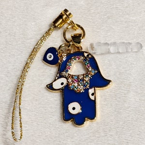 Könnte beinhalten: Ein goldener Anhänger mit einer blauen Hamsa-Hand, einem blauen Glücksbringer-Auge und einem klaren Kristall-Anhänger. Der Anhänger ist an einer goldenen Kette befestigt.
