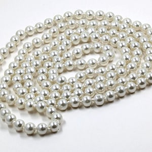 Peut inclure: Un long collier de fausses perles, chacune avec un éclat blanc cassé. Les perles sont enfilées ensemble, créant un collier continu et élégant. Le collier est présenté sur un fond blanc.