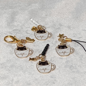 May include: Four gold-tone keychains with black cat charms inside white coffee cups. Each keychain has a different design and style.
