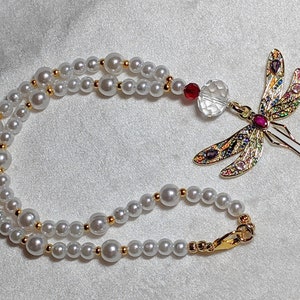 May include: A white pearl necklace with a gold dragonfly pendant. The dragonfly has colorful wings with rhinestones and a red gem in the center.