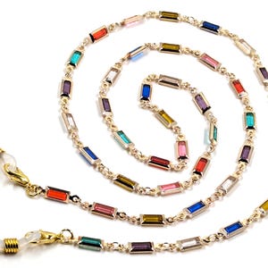 May include: A gold-toned eyeglass chain with rectangular, colorful gemstones. The chain features a variety of colors, including red, blue, green, and purple. The chain has gold-colored clasps on each end for attaching to glasses.