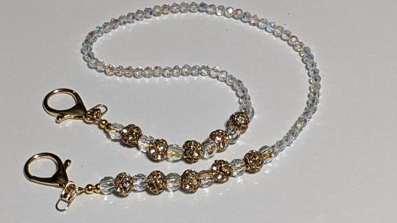 May include: A gold chain with clear and gold beads, featuring two gold clasps. The chain is designed to hold a mask or glasses.