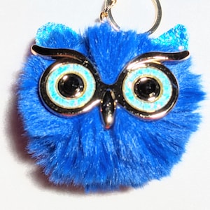 Pode incluir: Um chaveiro de coruja azul peludo com detalhes de glitter dourado e um anel de metal dourado. A coruja tem grandes olhos pretos com um anel azul e dourado ao redor.