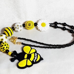 May include: A black beaded necklace with a yellow and black bee charm, yellow and white sunflower beads, and a white daisy charm.