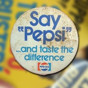 May include: Vintage Pepsi-Cola button with the text "Say 'Pepsi' and taste the difference." The button is white with blue text and a Pepsi-Cola logo at the bottom. The button has a worn, aged appearance.