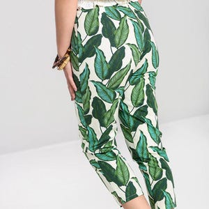 May include: Cropped trousers featuring a vibrant green and white tropical leaf pattern. The pants have a fitted silhouette, a high waist, and a straight leg cut that ends above the ankle, showcasing a bold botanical print on a light background.