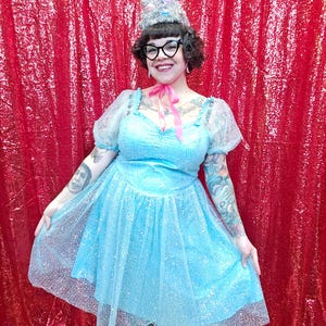 May include: A light blue dress with a glittery overlay, featuring short, puffy sleeves and a sweetheart neckline. The dress is paired with a silver, cone-shaped headpiece and black-framed glasses. A pink ribbon is tied around the neck.