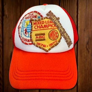 May include: A red and white trucker hat with embroidered patches. The patches include text that reads "2014 Bayern M", "Mixed League Champion", "Junior Aide", and "ABC 1976-1977". The hat has a red brim and a white mesh back.