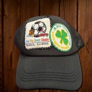 May include: Black trucker hat with mesh back and a curved brim. The hat features two embroidered patches: one with a soccer ball and text "River City Soccer Classic Peoria, Illinois", and another with a green shamrock and text "Jr. Irish Travel Soccer South Bend Indiana".