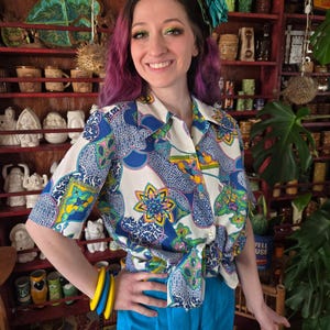 May include: A woman wearing a white button-down shirt with a blue, green, and yellow floral pattern, paired with blue shorts. She is also wearing yellow and blue bracelets. The background features shelves filled with various decorative items.
