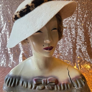 May include: A cream-colored hat with a wide brim and a dark brown and black leopard print band around the crown. The hat is displayed on a mannequin bust featuring a painted face with red lips and a sculpted bow detail on the chest.