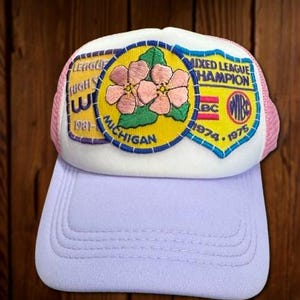 May include: A vintage trucker hat with a white front panel and a light purple brim. The hat features a patch with pink flowers, green leaves, and text that reads "Mixed League Champion" and "Michigan". The hat has a pink mesh back.