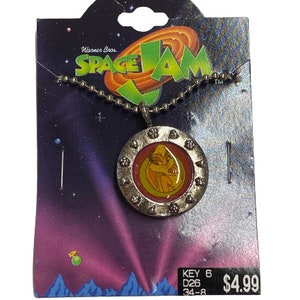 May include: A silver chain necklace with a round pendant featuring a cartoon character from the movie Space Jam. The pendant has a red and yellow background with a silver border. The pendant is on a card with the words "Warner Bros. Space Jam" and "KEY 6 D26 34-8 $4.99".