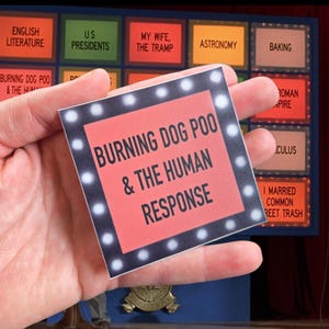 May include: A square sticker with the text "BURNING DOG POO & THE HUMAN RESPONSE" in white on a red background, framed by a border of white lights. The sticker is held in a hand, with other text blocks visible in the background.