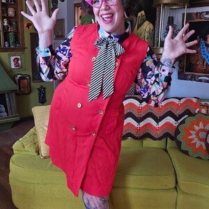 May include: A bright red, double-breasted dress with gold buttons. The dress is worn over a patterned blouse with a black and white striped tie. The person is wearing pink glasses and has tattoos on their legs.