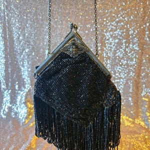 May include: A black beaded handbag featuring a triangular frame with a silver-toned metal clasp and an intricate filigree detail. The body of the bag is covered in small black beads and finished with a long fringe of black beaded strands.