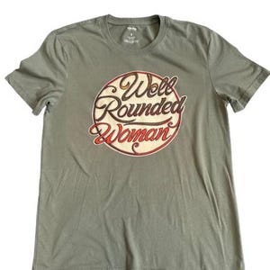 May include: Olive green t-shirt with a crew neck. The shirt features a circular graphic with the words "Well Rounded Woman" in a vintage-style font, with red and brown accents. The shirt is made of a soft material.