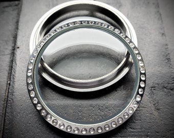 May include: A silver-toned locket with a clear glass center and a sparkling, gem-studded bezel. The locket is open, revealing the interior, and sits on a dark, textured surface. The design is elegant and suitable for personalization.