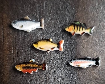 Puede incluir: Cinco figuritas de peces de colores sobre un fondo marrón oscuro. Los peces están hechos de cerámica y tienen detalles realistas.