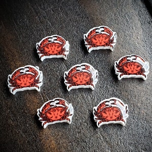 May include: Eight white ceramic crab-shaped buttons with red and black details. The buttons are arranged in a circle on a dark brown wooden surface.