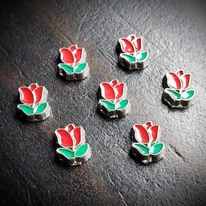 May include: Small, decorative charms in the shape of tulips. Each charm features a red flower with green leaves and a silver-colored border. The charms are scattered on a dark, textured surface.