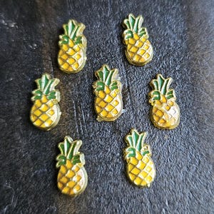 May include: Seven small, yellow pineapple charms with green tops and gold outlines are arranged on a dark surface. The charms have a textured appearance, and the green tops are detailed with leaf-like shapes.