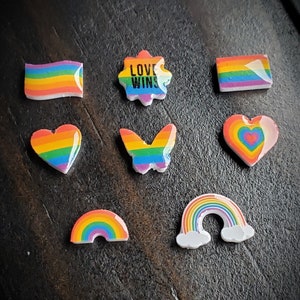 May include: A collection of colorful Pride-themed embellishments. The assortment includes a rainbow flag, a star with "LOVE WINS" text, hearts, a butterfly, and rainbows. The items feature vibrant rainbow colors and are set against a dark background.