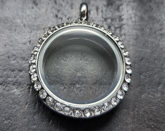 May include: A circular silver-toned floating locket pendant featuring a clear glass center framed by a border of small, clear, sparkling rhinestones. The locket has a small metal bail at the top for attachment to a chain or necklace cord.