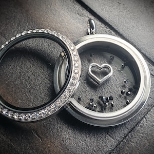May include: A silver locket with a clear glass insert and a silver heart charm inside. The locket is decorated with small clear rhinestones around the edge. The locket is open and the inside is filled with black glitter.