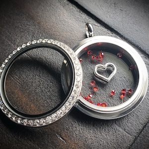 May include: Silver locket with a clear glass insert and a silver heart charm. The locket is decorated with small, clear rhinestones around the edge. The glass insert is filled with small, red rhinestones.