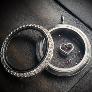 May include: A silver locket with a heart charm and a clear glass insert filled with small pink rhinestones. The locket is surrounded by a silver band with small clear rhinestones.