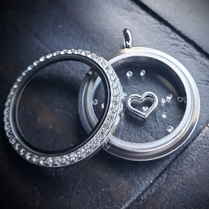 May include: Silver locket with a clear glass insert and a small heart charm inside. The locket is decorated with a row of clear rhinestones around the edge.