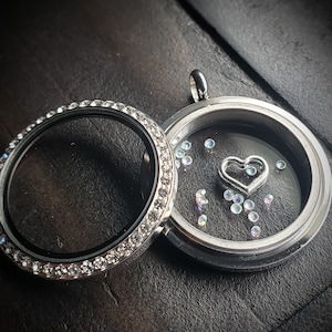 May include: A silver locket with a clear glass insert and a silver heart charm. The locket is surrounded by a row of clear rhinestones.