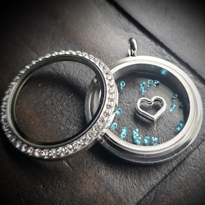 May include: A silver locket necklace with a clear glass insert and a small silver heart charm. The locket is surrounded by a row of clear rhinestones.